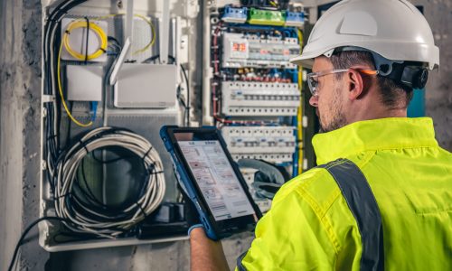 Man, an electrical technician working in a switchboard with fuses. Installation and connection of electrical equipment. Professional uses a tablet.